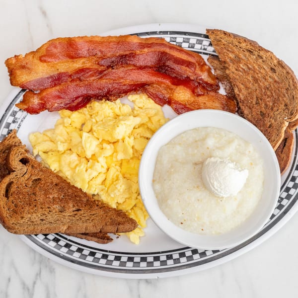 A diner breakfast with crispy bacon, scrambled eggs, grits with butter, and wheat toast.