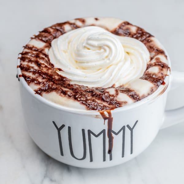Hot chocolate with whipped cream and chocolate drizzle in a white mug that says "YUMMY".